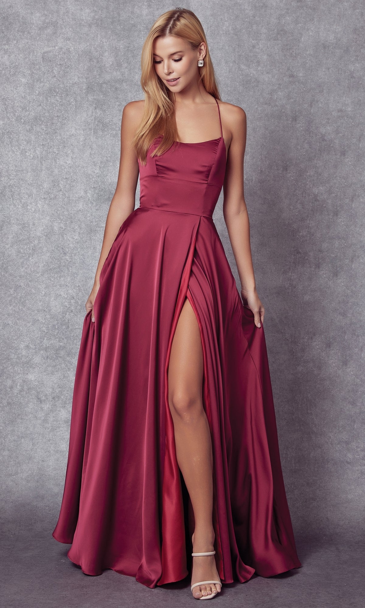 A woman wears the Strappy-Back Simple Long A-Line Prom Dress 278, a sleeveless, floor-length burgundy satin gown with a high slit, standing against a gray textured background.